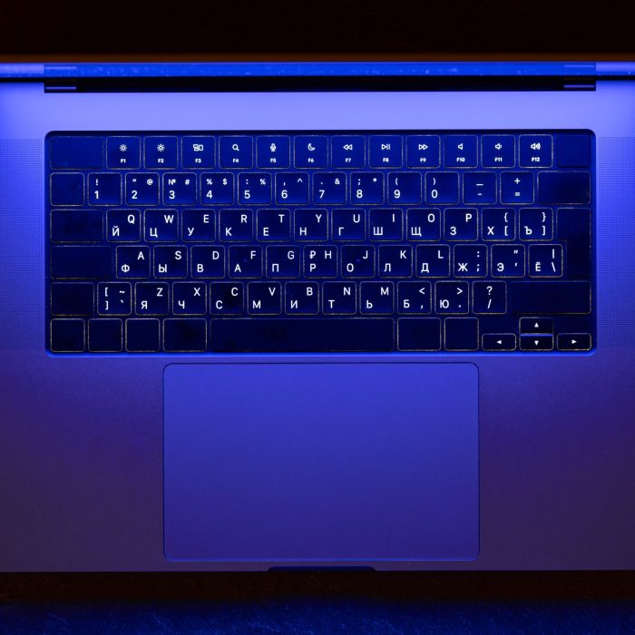 Overhead view of a laptop with Cyrillic keyboard glowing in deep blue neon light. Creates a focused night workspace mood for coding design and other digital projects.
